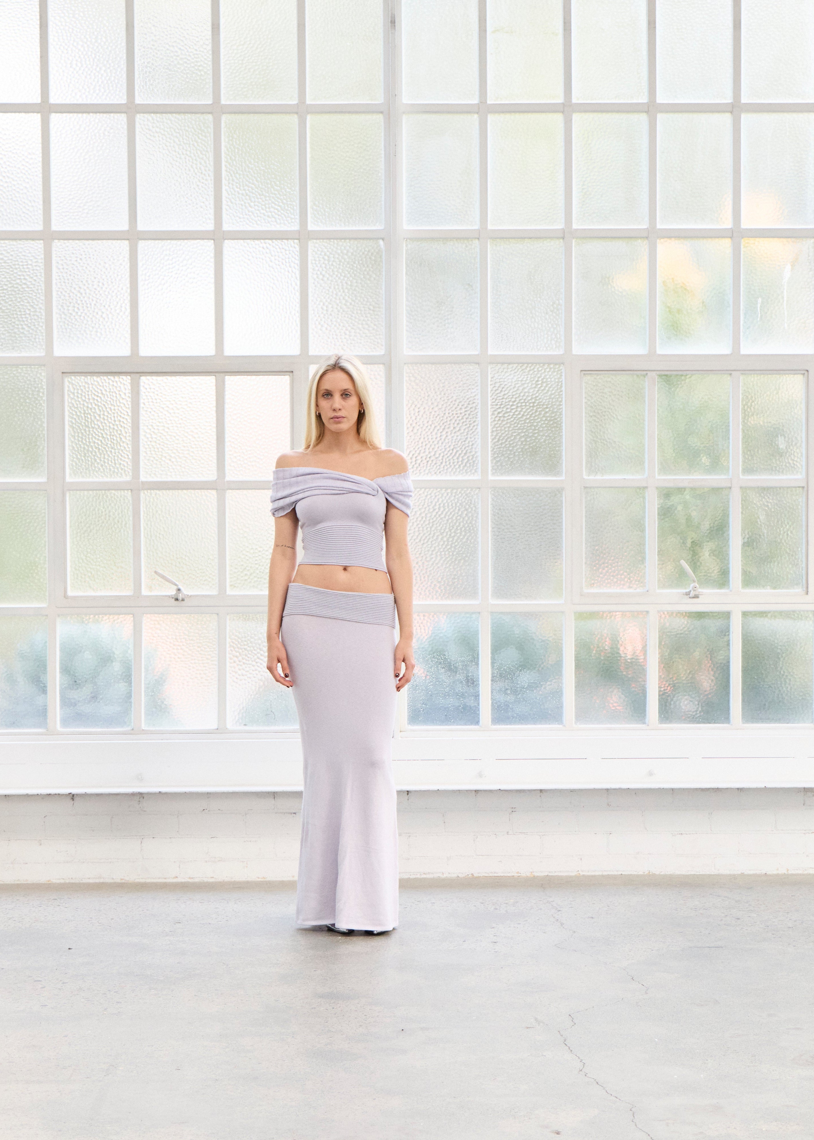 Woman in wisteria knitwear top and skirt standing in front of window